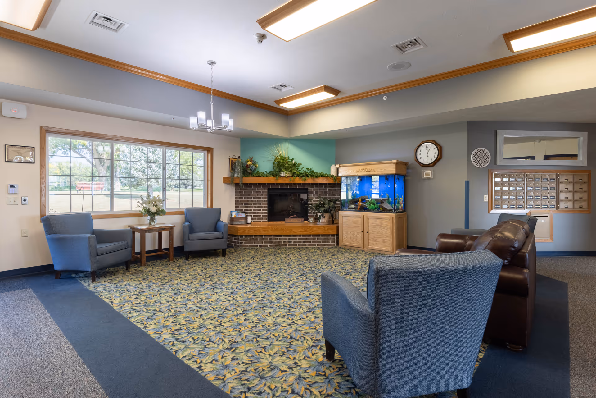 A cozy senior living facility common area with blue upholstered chairs and a brown leather sofa arranged around a patterned carpet. There is a brick fireplace with a wooden mantle decorated with plants, and a large fish tank on a wooden cabinet next to it. A large window lets in natural light, and a wall clock and mailboxes are visible on the wall.