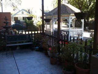 Shaded covered patio with potted plants and a bench overlooking a small fenced yard and a white gazebo.