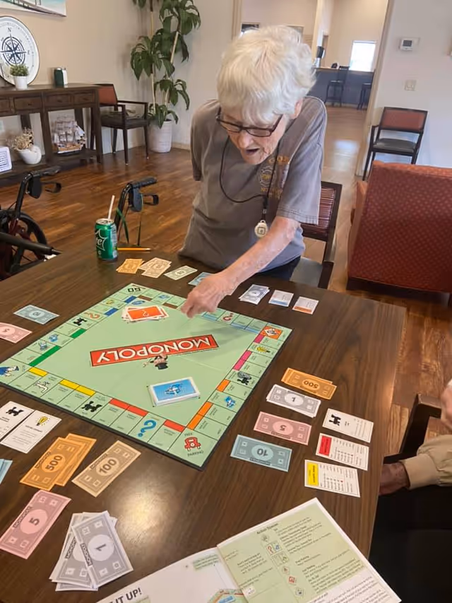 An elderly woman with white hair and glasses is sitting at a wooden table playing a game of Monopoly. The table is covered with Monopoly money, cards, and game pieces. The room has wooden floors, chairs, a plant, and a decorative shelf in the background.