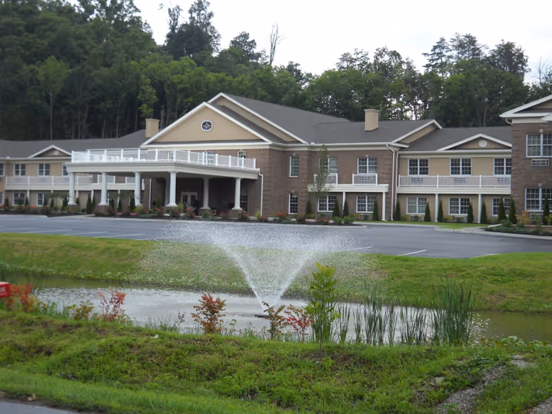 Exterior view of Azalea Hills Assisted Living facility showing a large two-story building with beige and brick facade, a covered entrance with white railings, surrounded by trees and greenery. In the foreground, there is a small pond with a water fountain spraying water upwards.