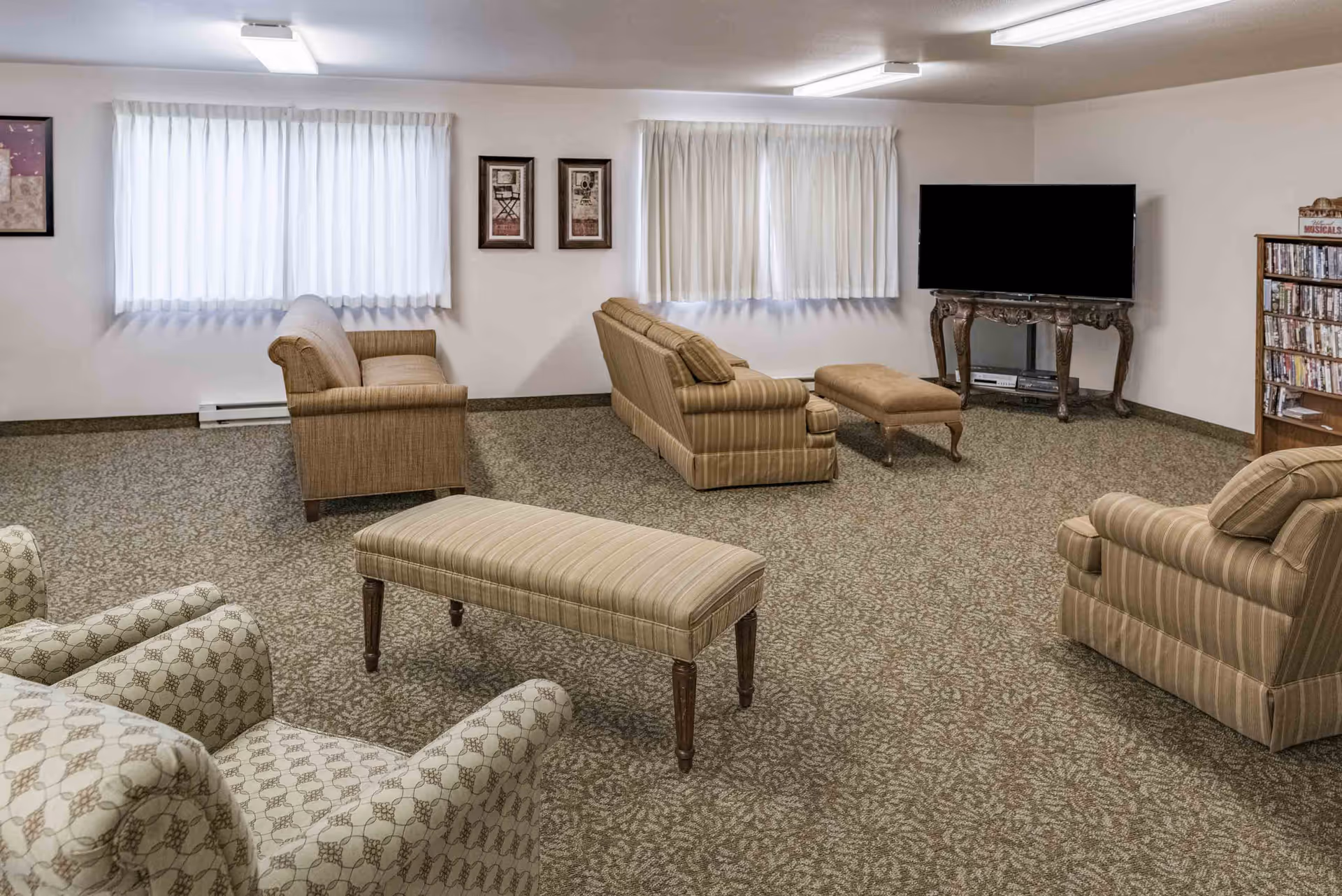 Spacious common living room with upholstered chairs and sofas, ottomans, a TV on a stand, patterned carpet, and windows with sheer curtains.