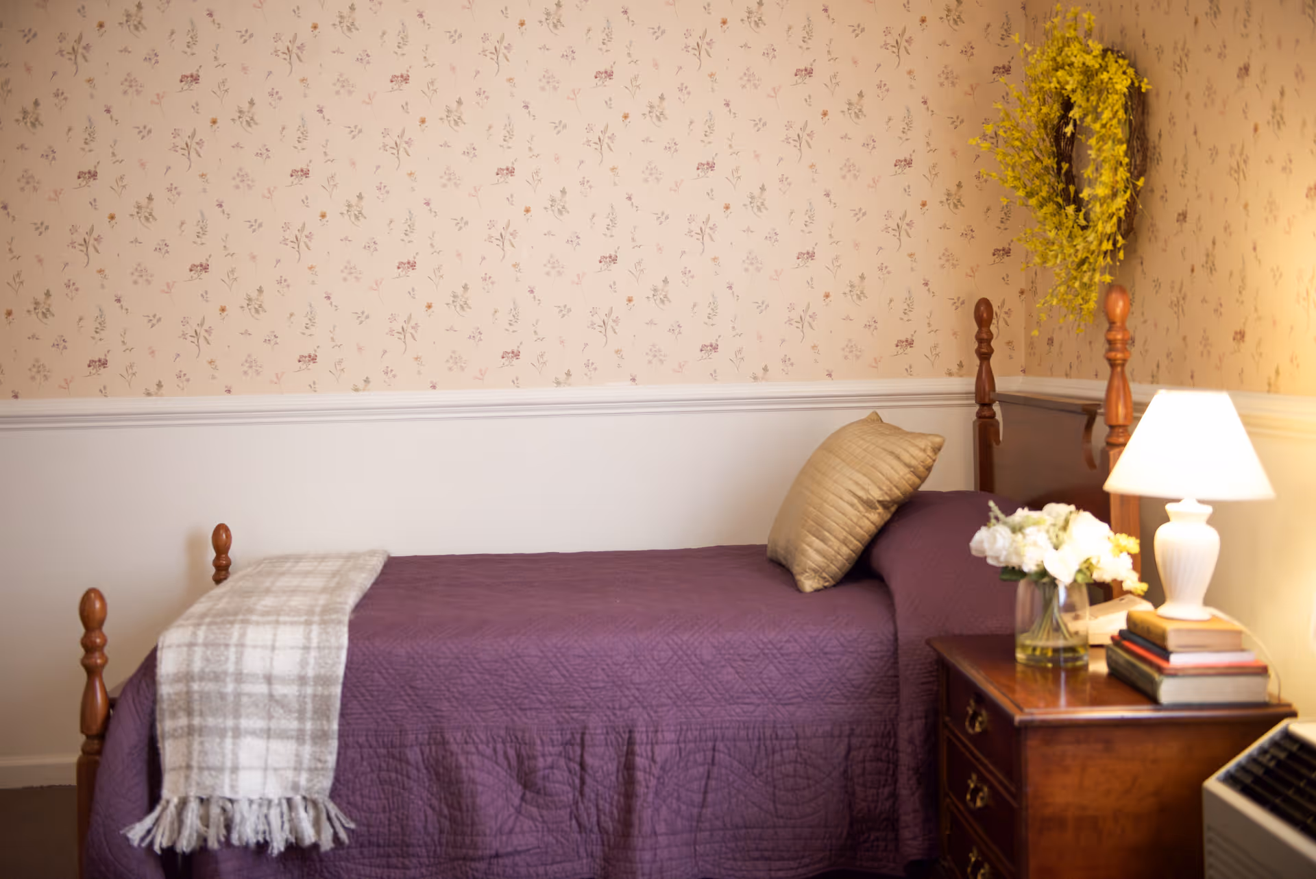 A cozy bedroom with a single wooden bed covered in a purple quilt and a beige pillow. A plaid throw blanket is draped over the foot of the bed. Next to the bed is a wooden nightstand with a white lamp, a vase of white flowers, and a small stack of books. The wall behind the bed has floral wallpaper with a yellow wreath hanging above the headboard.