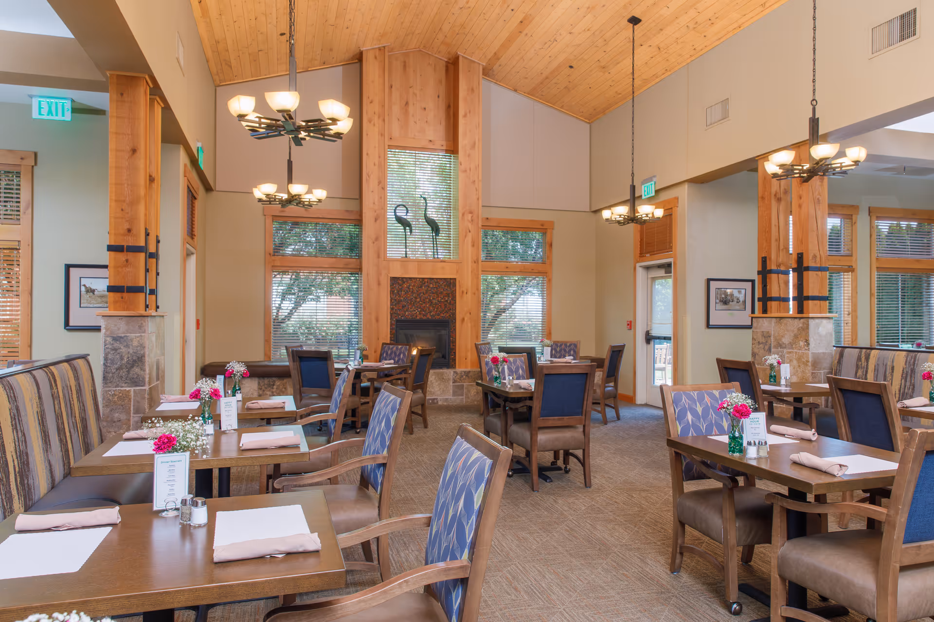 A bright and spacious dining room with wooden tables and chairs, some with patterned upholstery. Tables are set with placemats, napkins, and small flower arrangements. The room features large windows with wooden blinds, a high wooden ceiling, and decorative light fixtures. There is a fireplace with two bird sculptures on the mantel and exit signs above doors.