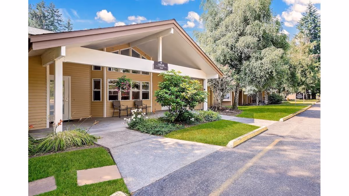 Exterior view of a senior living facility building with a covered entrance, beige siding, and a small garden area with green grass and shrubs. There are two chairs on the porch and a sign above the entrance that reads 'Cedar'. Trees and a clear blue sky with some clouds are visible in the background.