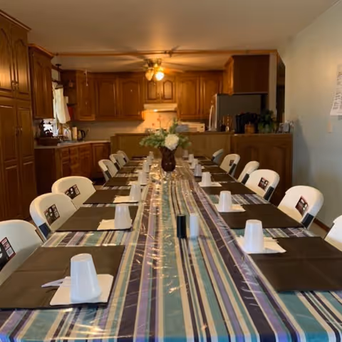 Long communal dining table set with placemats, disposable cups, and a floral centerpiece in a dining area with a kitchen in the background.