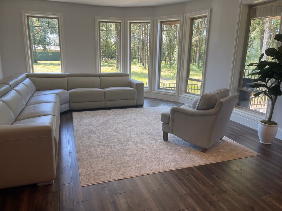 Bright living room with a large beige sectional sofa, an armchair on a light area rug, and tall windows overlooking trees.