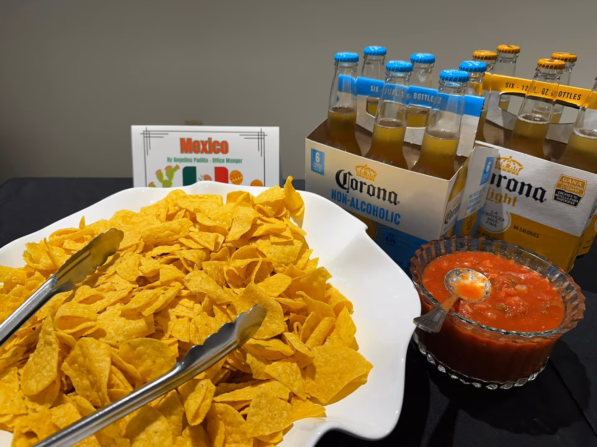A white bowl filled with yellow corn chips with metal tongs resting on top, next to a glass bowl of red salsa with a spoon. Behind the bowls are two six-packs of Corona beer, one labeled non-alcoholic and the other light. A small sign in the background reads 'Mexico' with a Mexican flag and illustrations of a cactus and maracas.