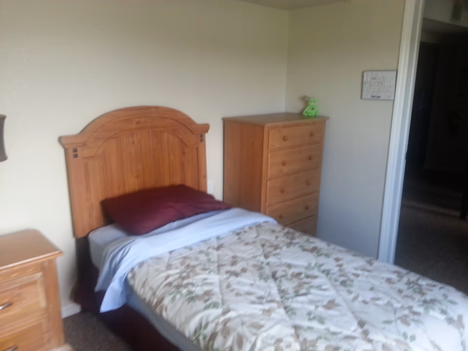 A small bedroom with a single wooden bed featuring a floral patterned comforter and a burgundy pillow. Next to the bed is a wooden nightstand, and against the wall is a wooden chest of drawers with a small green stuffed toy on top. A floor plan is mounted on the wall near the doorway.