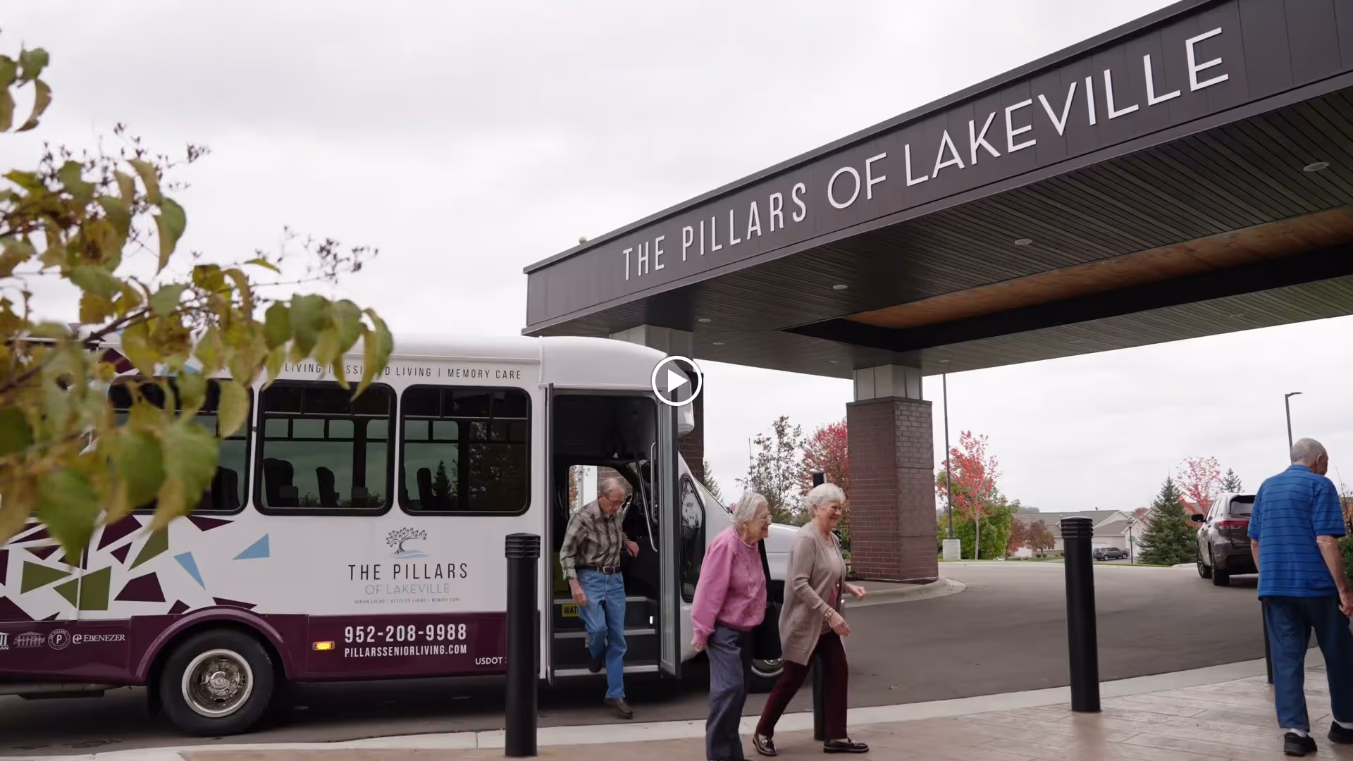 The entrance area of The Pillars of Lakeville senior living facility with a shuttle bus parked nearby. Several elderly people are walking near the entrance under a large sign displaying the facility's name.