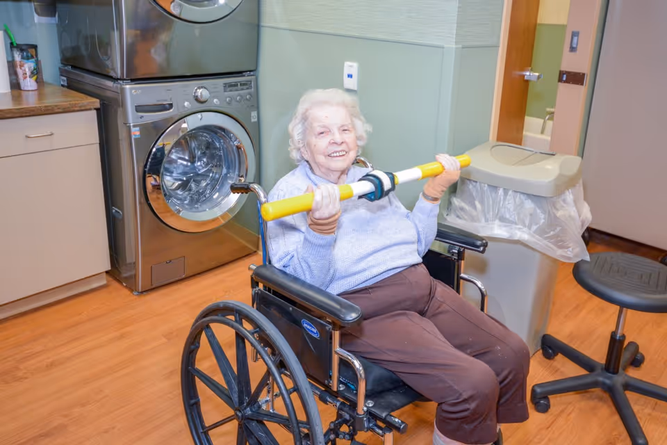 An elderly woman in a wheelchair holding a yellow and white exercise bar, smiling in a room with a stacked washer and dryer, a trash can, and a rolling stool.