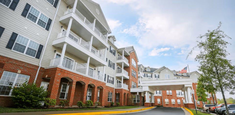 Exterior view of a multi-story senior living facility building with brick and light-colored siding, balconies, an entrance canopy, and an American flag on a flagpole. There are some trees and parked cars visible near the entrance.