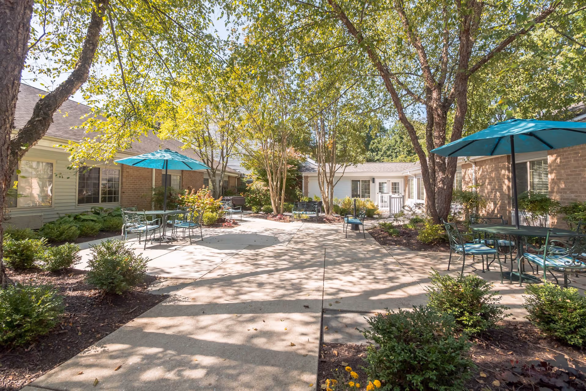 Outdoor courtyard area at Lighthouse Senior Living at Ellicott City featuring paved walkways, green shrubs, trees providing shade, and several metal tables with teal umbrellas and chairs for seating.