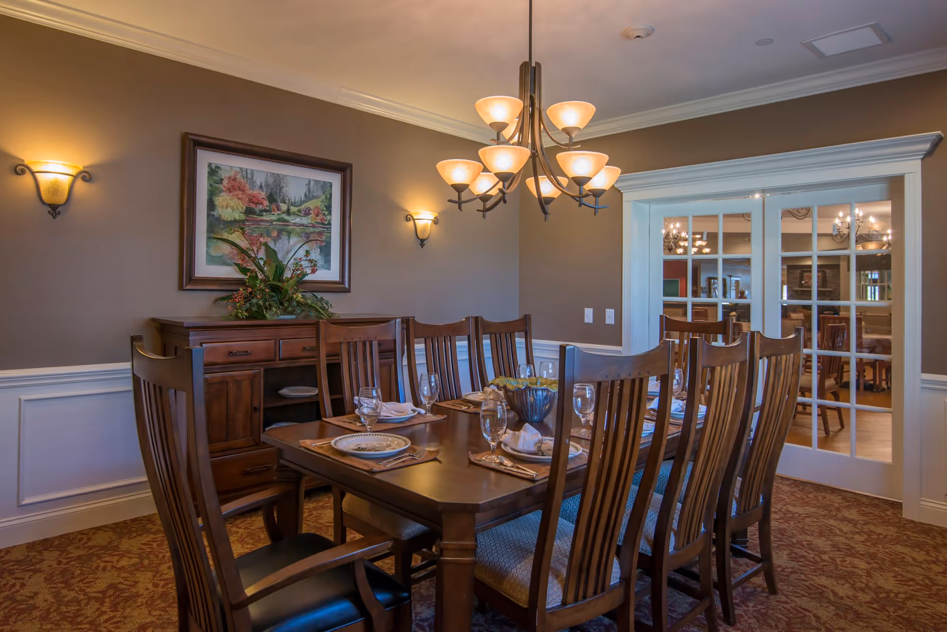 A formal dining room with a wooden dining table set for eight people. The table has plates, glasses, napkins, and silverware arranged neatly. There is a chandelier with multiple lights hanging above the table. On the wall, there are two wall sconces and a framed painting of a scenic landscape. A wooden sideboard with decorative plants is against the wall. The room has beige walls with white wainscoting and a patterned carpet. Glass-paneled double doors lead to another room.