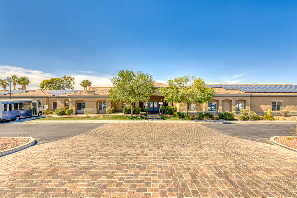 Front exterior view of a single-story senior living facility building with a paved driveway, green trees, shrubs, and a blue sky. A shuttle bus is parked on the left side of the building.