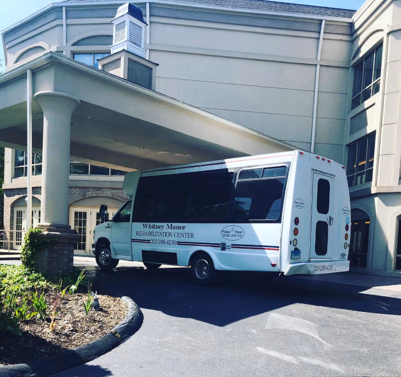 A Whitney Manor Rehabilitation Center shuttle bus parked under the covered entrance/portico of a beige multi-story building.