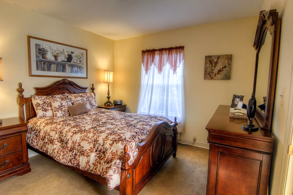 A cozy bedroom with a wooden bed frame and matching furniture including a nightstand and dresser with a mirror. The bed is made with a floral patterned comforter and pillows. There is a window with sheer curtains and a valance, a lamp on the nightstand, and framed artwork on the walls.