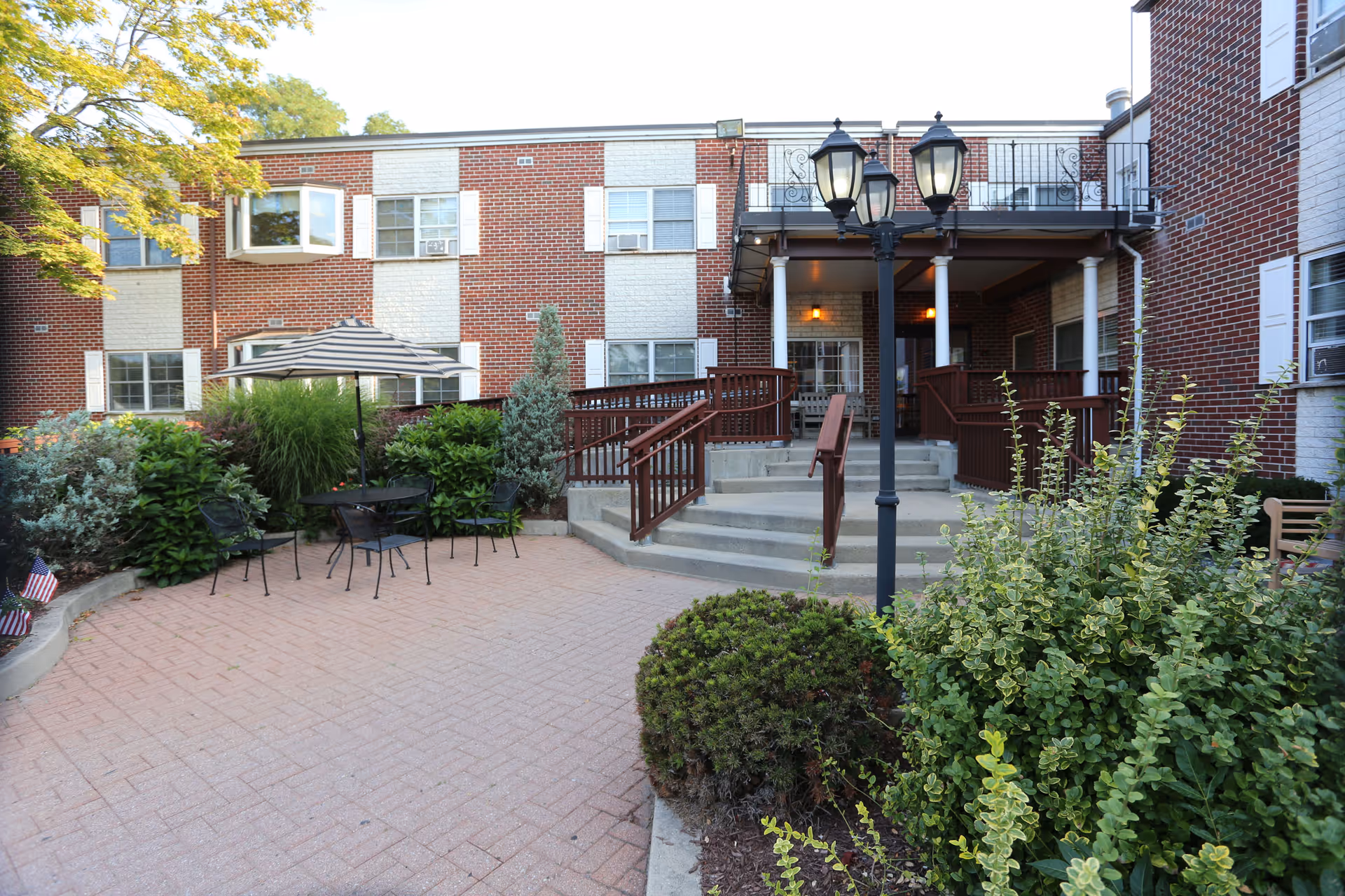 Outdoor courtyard area of a senior living facility with brick building in the background. The courtyard features a paved patio with a table, chairs, and an umbrella surrounded by various green shrubs and plants. There are steps and ramps leading to a covered porch with railings and outdoor lighting fixtures.
