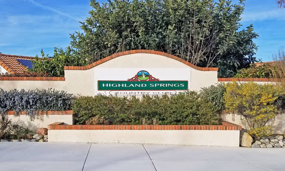 A landscaped stucco and brick entrance sign that reads "Highland Springs Country Club" with shrubs and trees behind it.