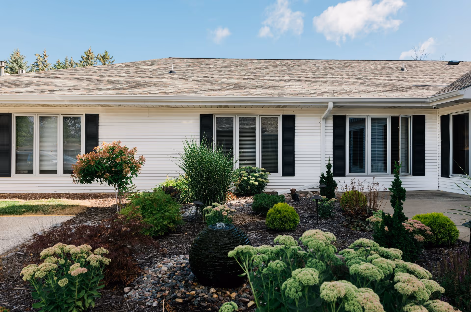 Exterior view of a single-story building with white siding and black window shutters. In front of the building is a landscaped garden with various green shrubs, plants, and a small water feature. The sky is clear with a few clouds.