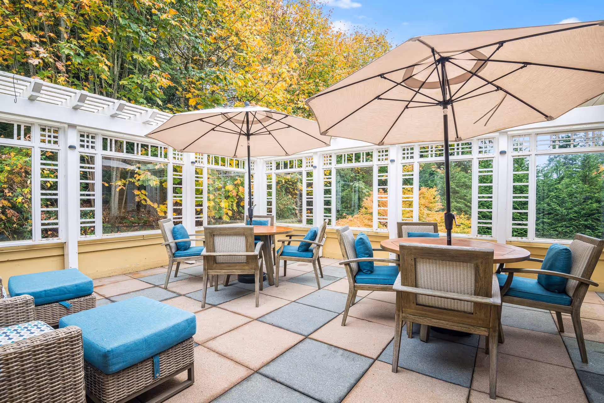 Sunlit enclosed patio with round tables, large umbrellas, wicker chairs and blue cushions overlooking autumn foliage.