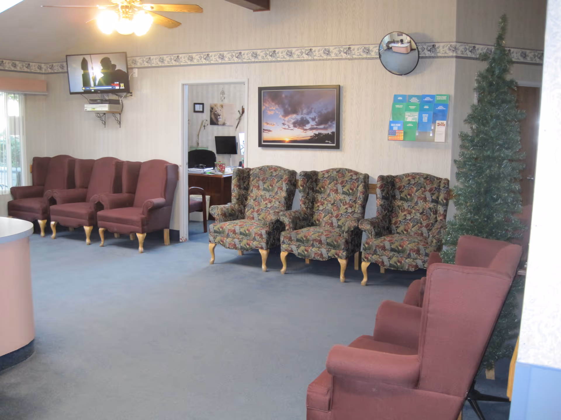 A waiting or common area with multiple upholstered armchairs in solid maroon and floral patterns arranged along the walls. A small artificial Christmas tree is placed near the right side. A wall-mounted TV is on the left side above the chairs, and a framed picture of a sunset hangs on the wall. There is a doorway leading to an office with a desk and computer visible. A circular convex mirror and a wall-mounted brochure holder are also visible on the wall.