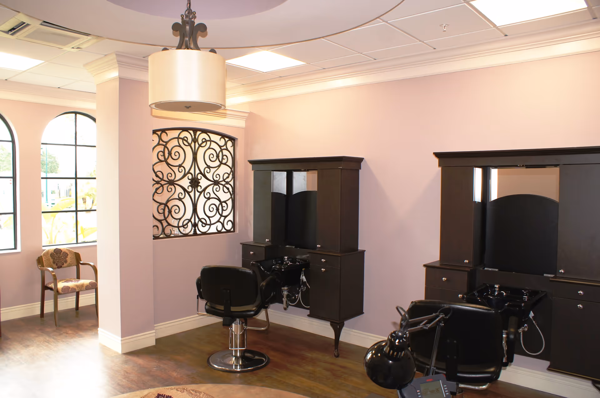 Interior view of a salon area with two black salon chairs in front of black cabinetry with mirrors and sinks. The room has light purple walls, wooden flooring, a decorative wrought iron window, and a beige patterned chair near large arched windows letting in natural light.
