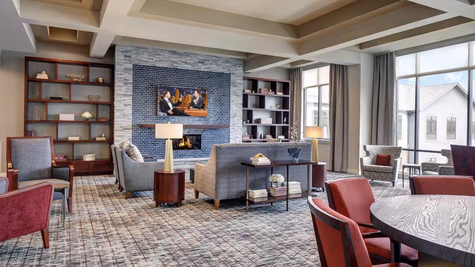 A spacious and well-lit living room area with multiple seating options including armchairs and sofas arranged around a modern fireplace with a mounted flat-screen TV. Large windows with curtains allow natural light to fill the room. Bookshelves with decorative items flank the fireplace, and a round table with red chairs is visible in the foreground.