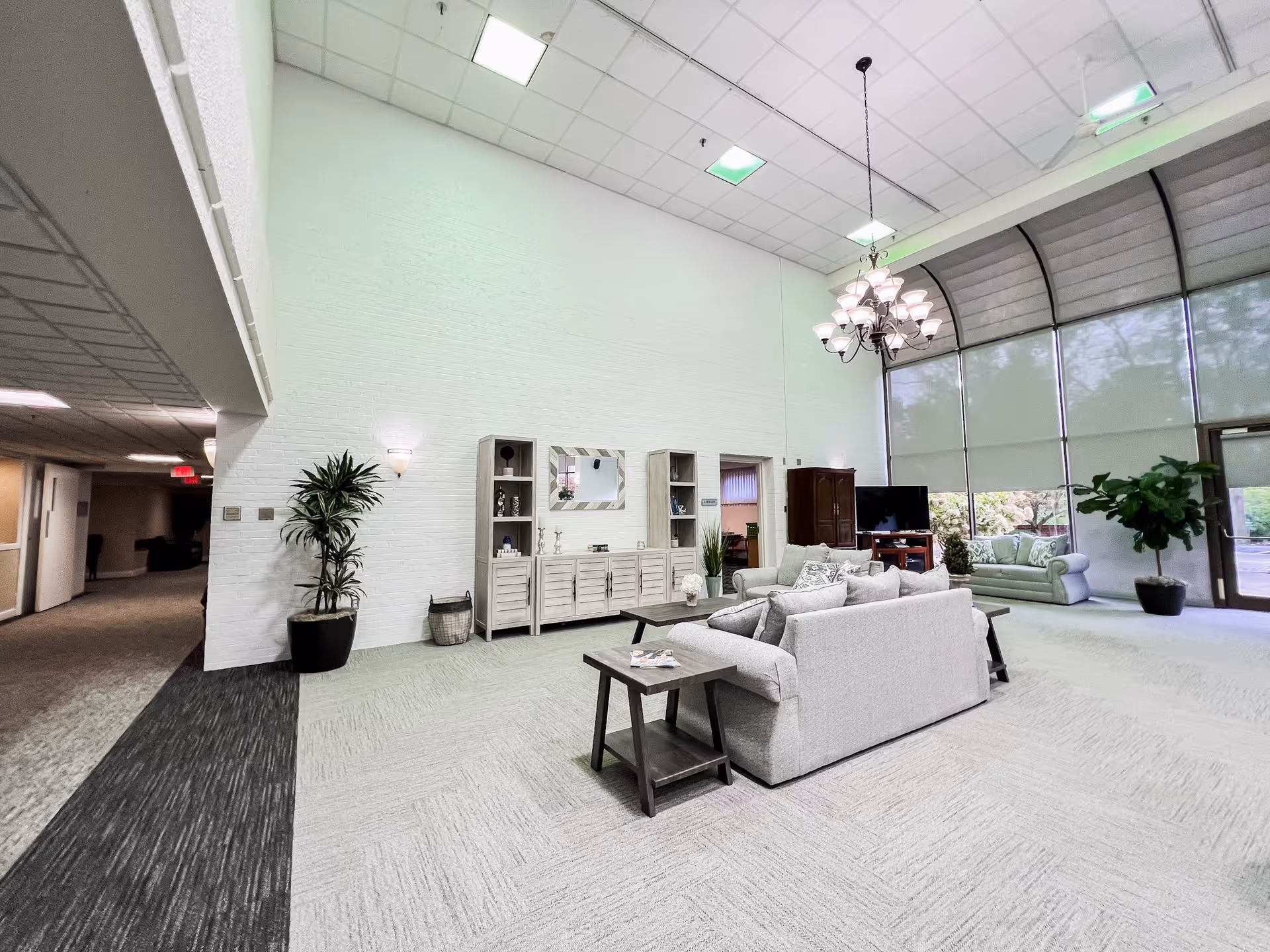 A spacious and well-lit living room area in The Trinity Community at Beavercreek featuring light gray sofas, a wooden coffee table, a TV on a stand, large windows with roller shades, a chandelier, potted plants, and a white brick wall with shelves and a mirror.