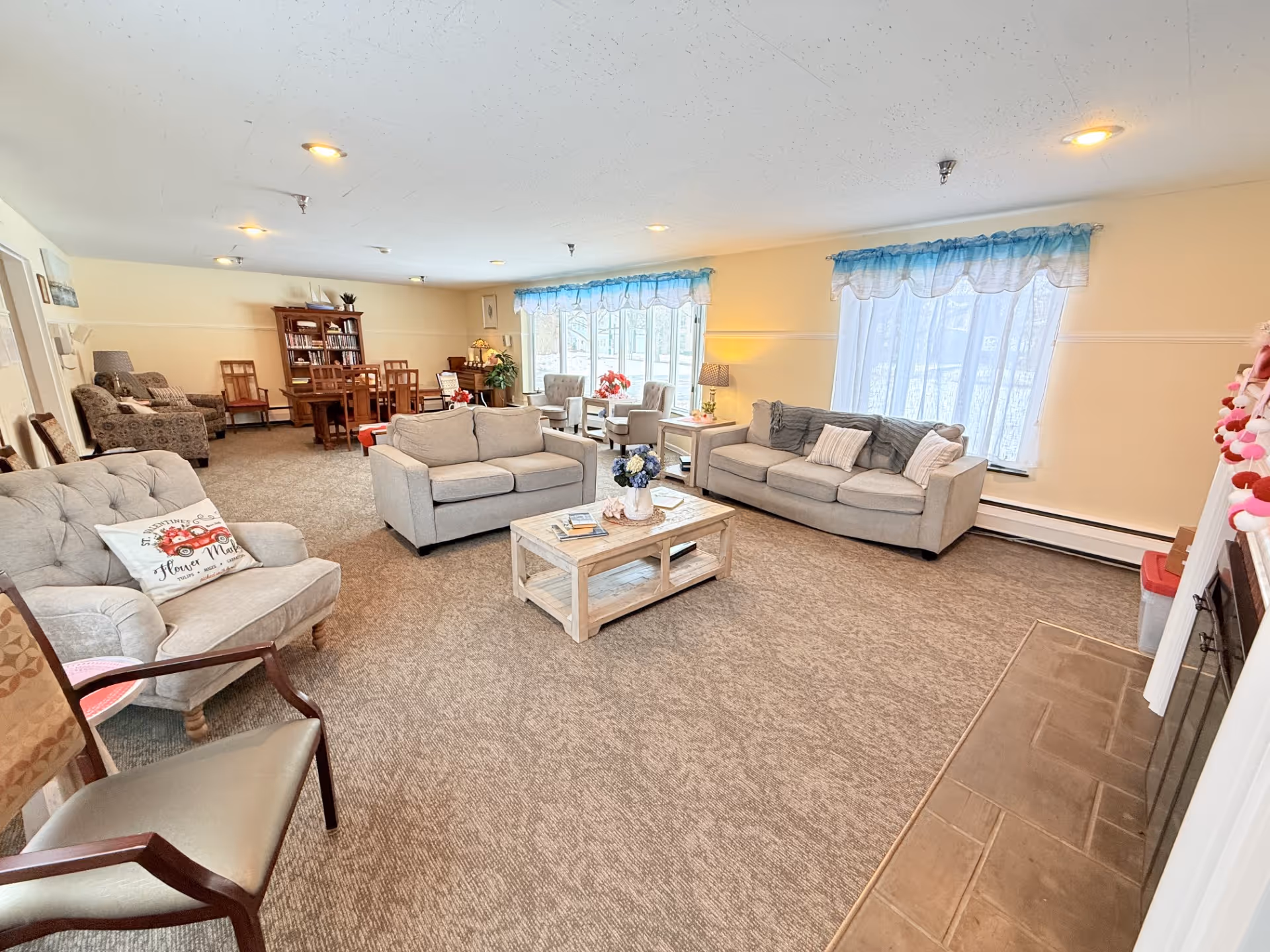 A spacious and well-lit living room in a senior living facility with multiple seating options including sofas, armchairs, and wooden chairs. The room features large windows with blue valances, a coffee table with decorative items, a fireplace on the right, and a dining area with a wooden table and chairs in the background. The carpeted floor and soft lighting create a cozy and inviting atmosphere.
