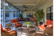 A screened-in porch area with wicker furniture including chairs and a loveseat with red cushions and decorative pillows. There is a round glass-top coffee table with a potted plant in the center. Ceiling fans are mounted on the covered roof, and large windows provide a view of the outdoor area.