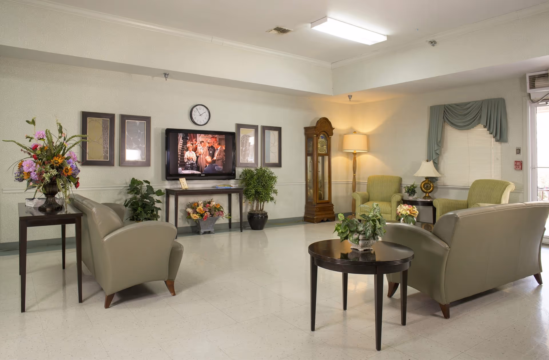 Well-lit senior living lounge with sofas and armchairs arranged around tables, a wall-mounted TV, grandfather clock and decorative plants.
