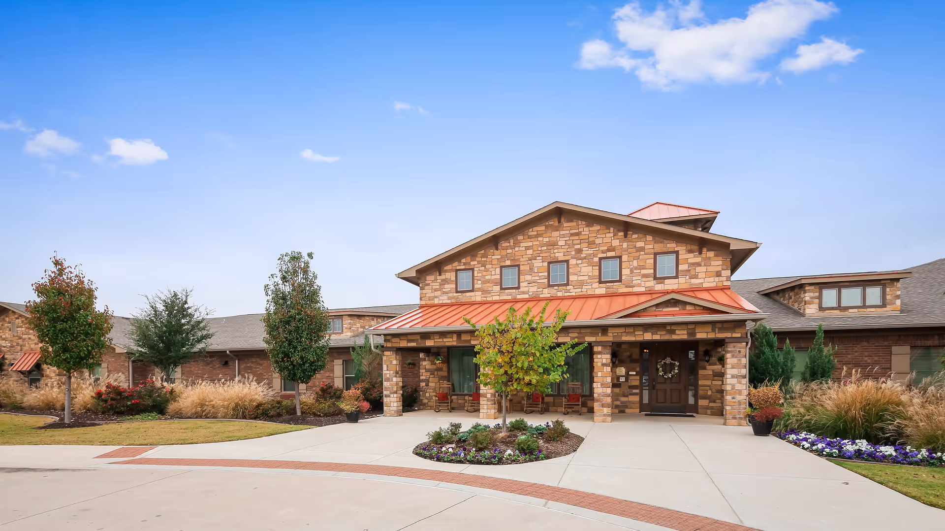 Front exterior view of Rosewood Assisted Living & Memory Care facility featuring a stone facade, red metal roof accents, a covered entrance with seating, landscaped garden beds, and a clear blue sky.