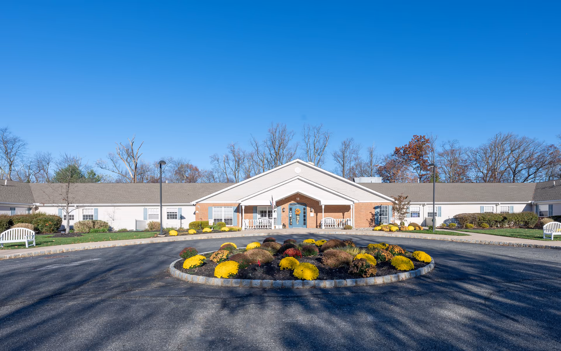 Front exterior view of Arden Courts - ProMedica Memory Care Community in Whippany, showing a single-story building with a central entrance, a circular driveway with a landscaped roundabout featuring yellow and red flowers, white benches on either side, and trees in the background under a clear blue sky.