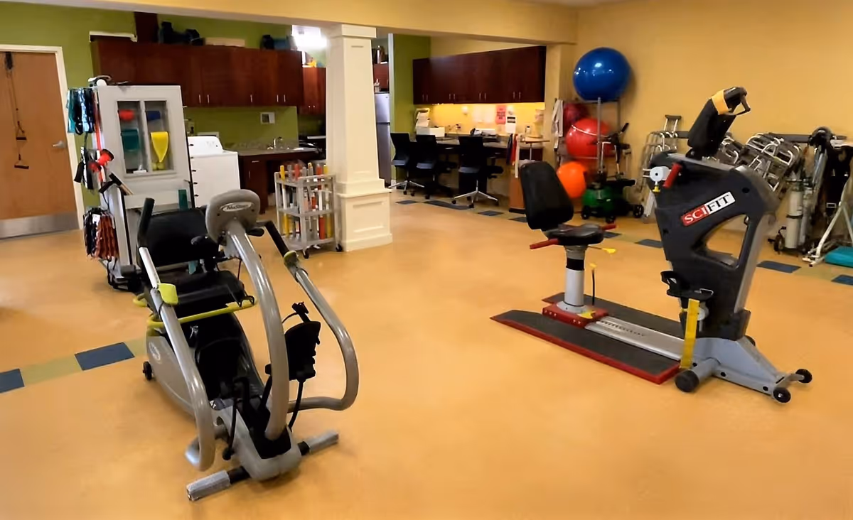 A fitness and rehabilitation room with exercise equipment including a recumbent bike and a SciFit machine. The room has a yellow floor, green and yellow walls, and a workspace with chairs and desks in the background. Various exercise balls and mobility aids are also visible.