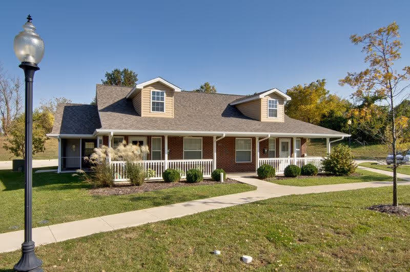 Single-story brick senior living building with a covered front porch, dormer windows, and a landscaped lawn under a clear blue sky.
