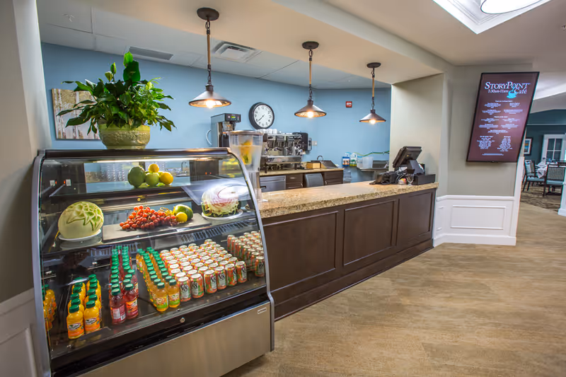 Interior view of a cafe counter at StoryPoint Chesterton with a glass display case containing bottled juices and decorative fruit carvings. Behind the counter are coffee machines, a clock on a blue wall, and three hanging pendant lights. A digital menu board is mounted on the wall to the right, and a seating area is visible in the background.
