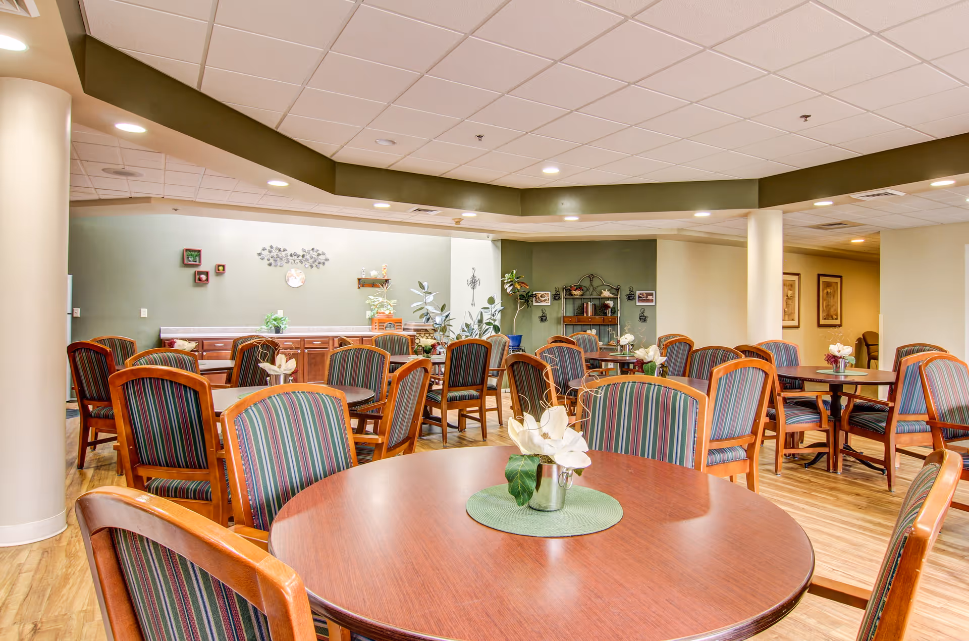 Communal dining room with round wooden tables and striped upholstered chairs, decorated with plants and wall art.
