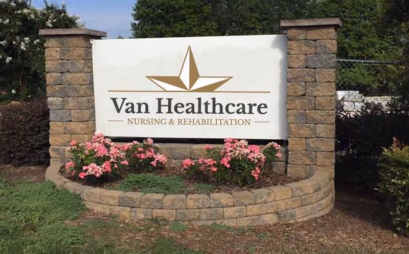 Outdoor sign for Van Healthcare Nursing & Rehabilitation, mounted on a stone structure with pink flowers planted in front and greenery in the background.