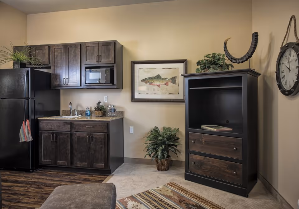Small kitchenette and living area with dark wood cabinets, black refrigerator and microwave, a sink and counter, framed fish artwork, potted plant, and a tall dark storage cabinet.