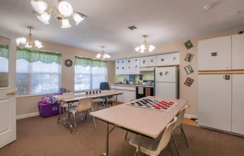 A bright room with two tables and chairs, one table has a checkerboard game set up. The room has large windows with green valances, white cabinets with coffee cup decorations, a refrigerator, and a stove. The walls have decorative letters spelling 'CAFE'. The carpet is brown, and there are ceiling light fixtures with multiple bulbs.
