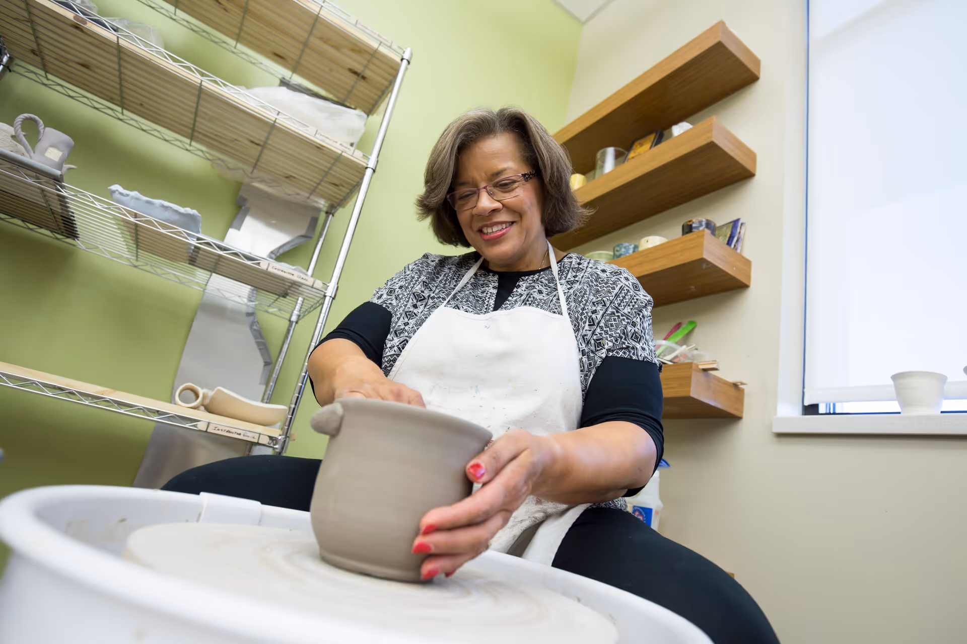 A woman wearing glasses and a white apron is shaping a clay pot on a pottery wheel in a room with green walls, metal shelves holding pottery items, and wooden wall shelves with various small objects.