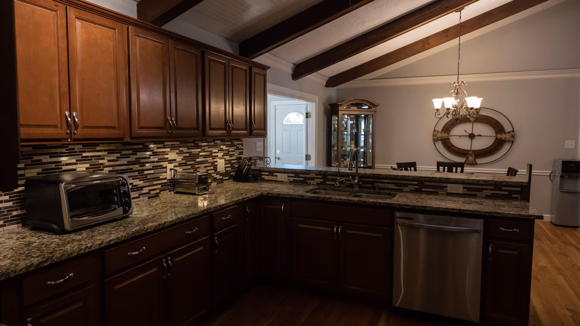 Open kitchen with dark wood cabinets, granite countertops, a mosaic tile backsplash, stainless steel appliances, and a view into a dining area.