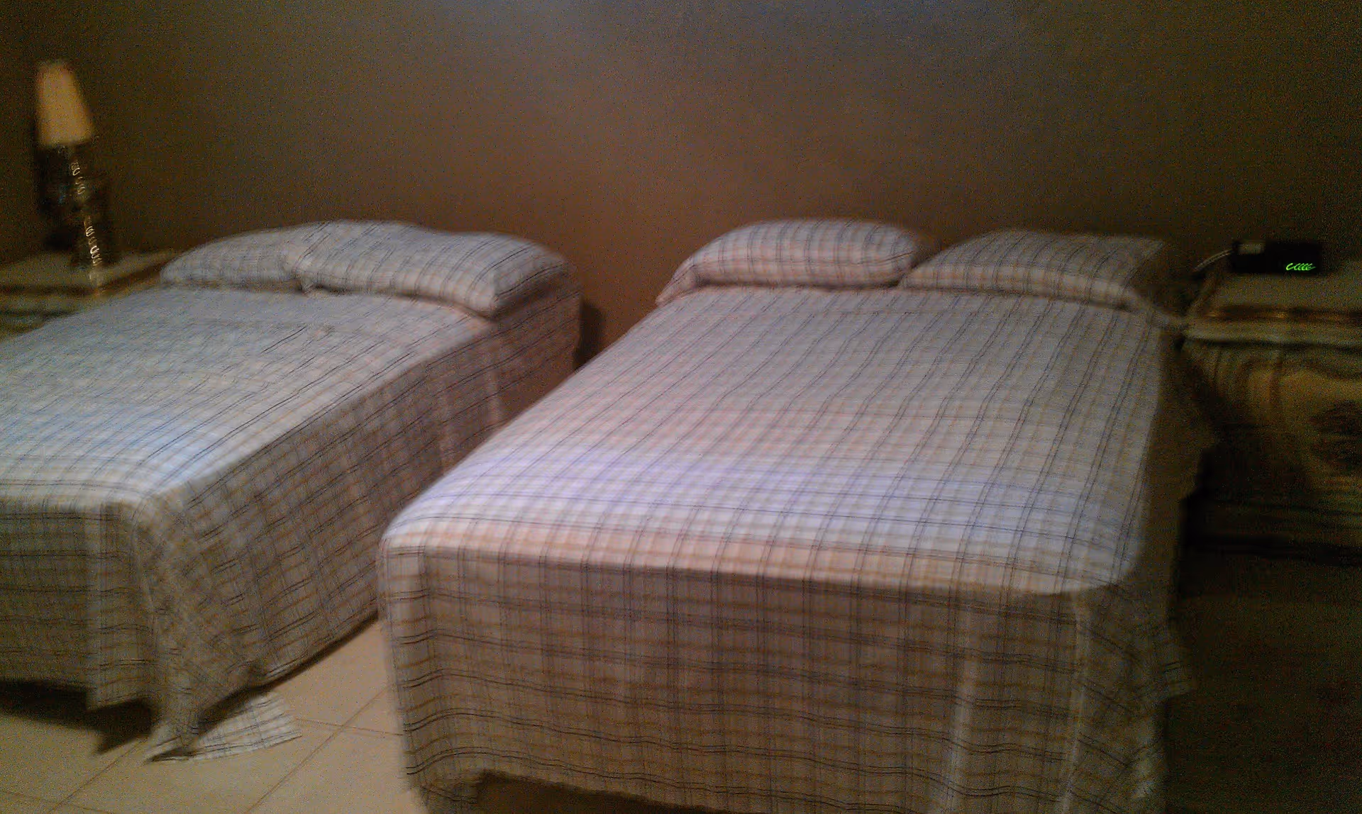 A bedroom with two beds side by side, each covered with plaid bedspreads and matching pillows. There is a nightstand between the beds with a lamp on the left side and a digital clock on the right side. The walls are painted brown and the floor is tiled.