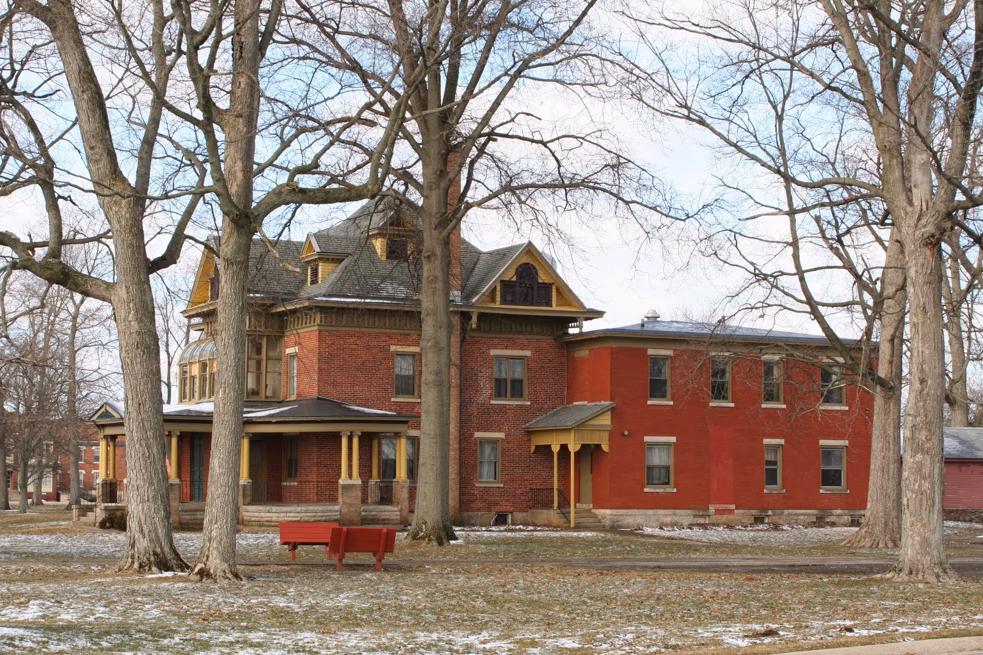 A large, historic brick building with yellow trim surrounded by leafless trees in a grassy area with patches of snow. There is a red bench in front of the building and a clear sky above.