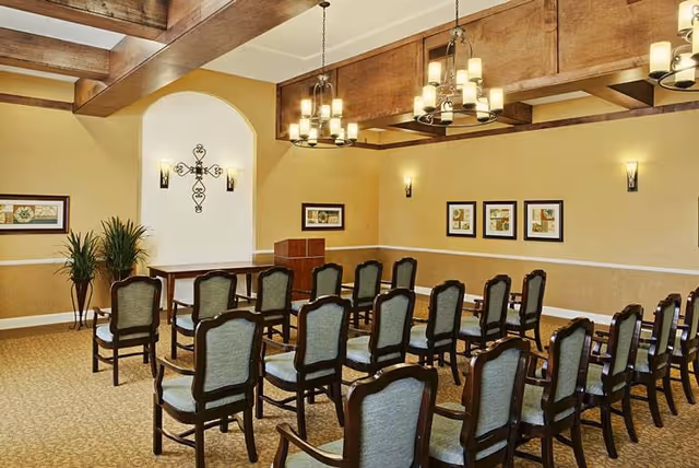 A well-lit room with rows of wooden chairs with cushioned seats arranged facing a small wooden podium and table. The walls are painted beige with white trim and decorated with framed pictures and a decorative metal cross. The ceiling features wooden beams and hanging light fixtures with multiple bulbs.
