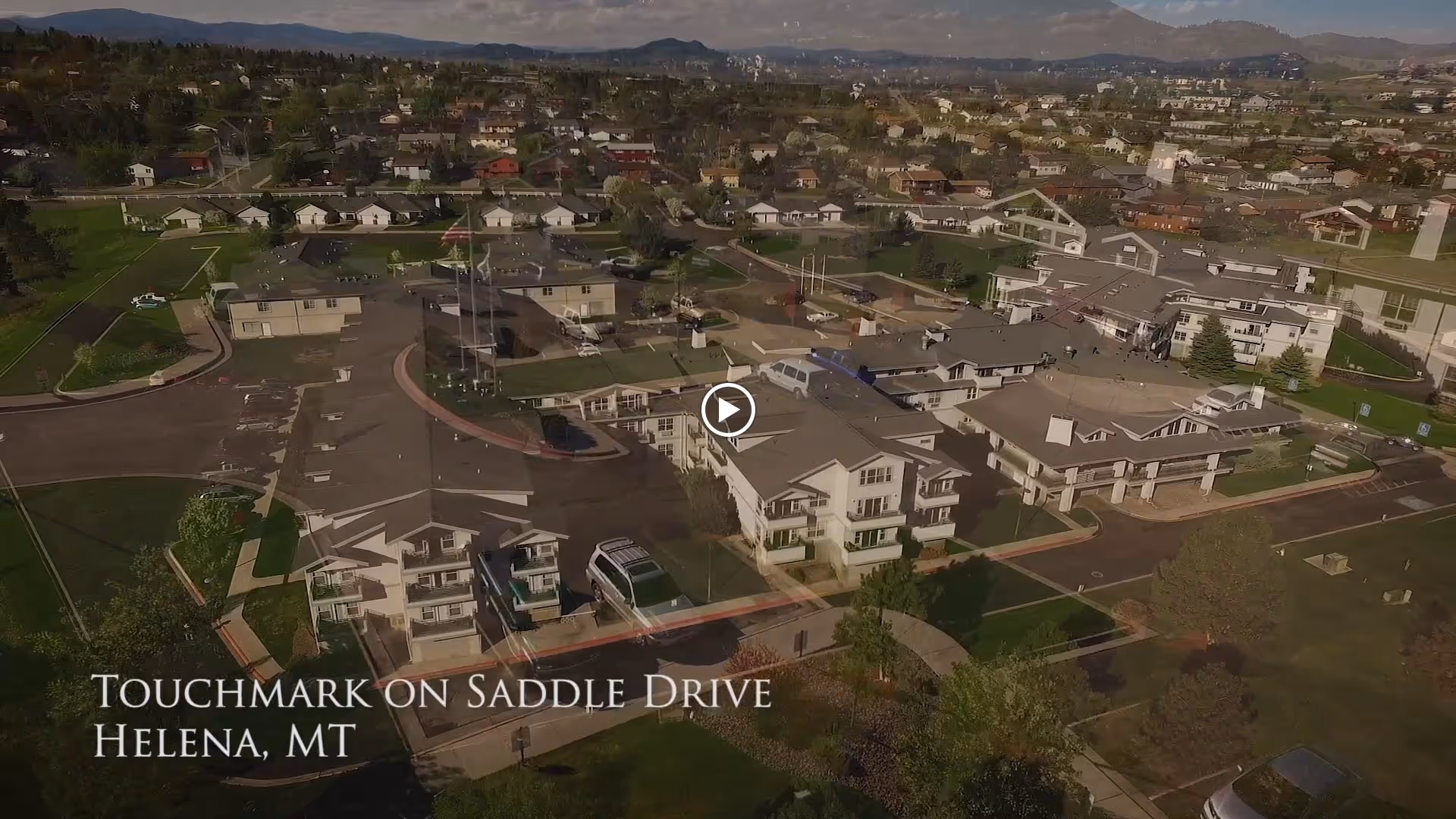 Aerial view of the Touchmark on Saddle Drive senior living facility in Helena, Montana, showing multiple buildings, parking areas, and surrounding neighborhood with hills in the background under a partly cloudy sky.