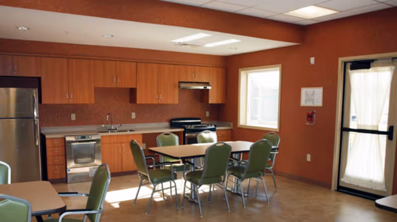 Communal dining area with several tables and green chairs in front of a fully equipped kitchen with wooden cabinets and stainless appliances.