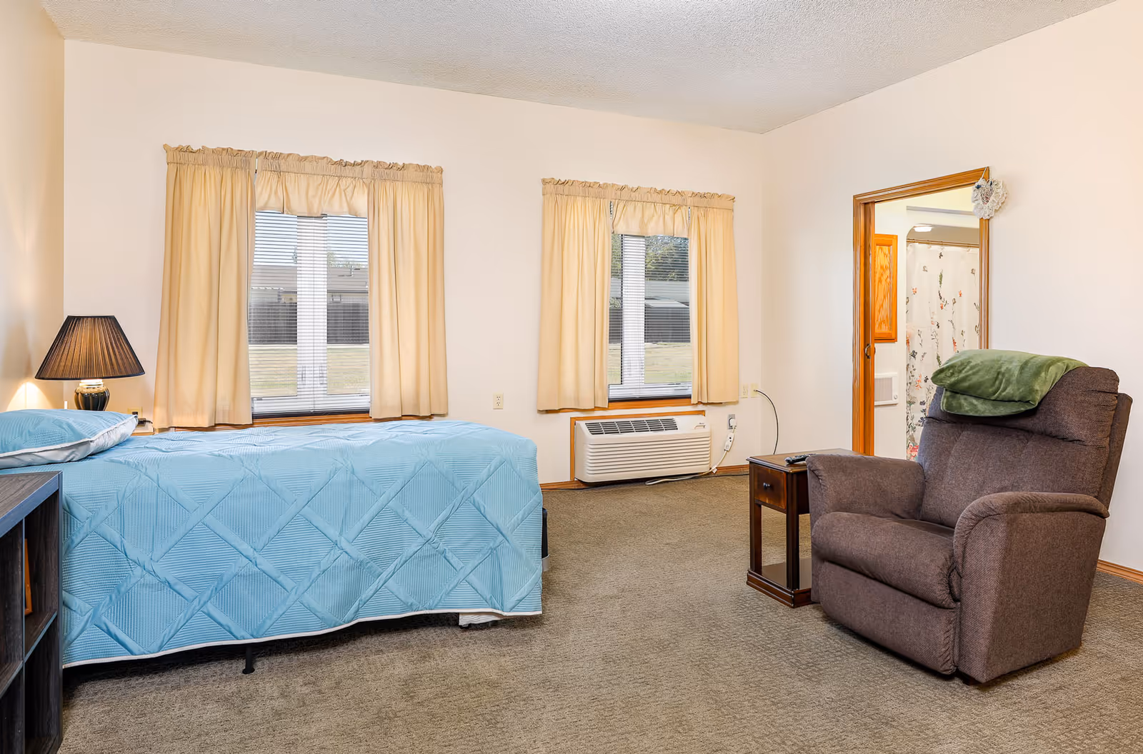 A bedroom in an assisted living facility featuring a single bed with a light blue quilt, a bedside table with a lamp, two windows with beige curtains, a brown recliner chair with a green blanket draped over the back, and a small wooden side table. A doorway leads to a bathroom with a shower curtain visible.
