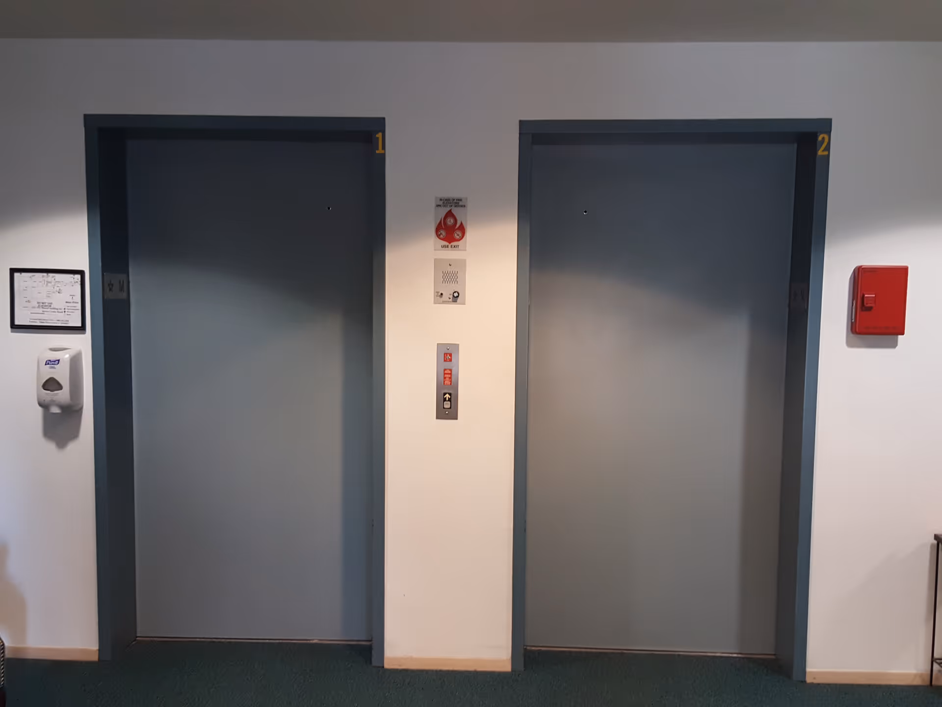 Two closed elevator doors numbered 1 and 2 in a hallway with a fire alarm pull station and a hand sanitizer dispenser mounted on the walls.
