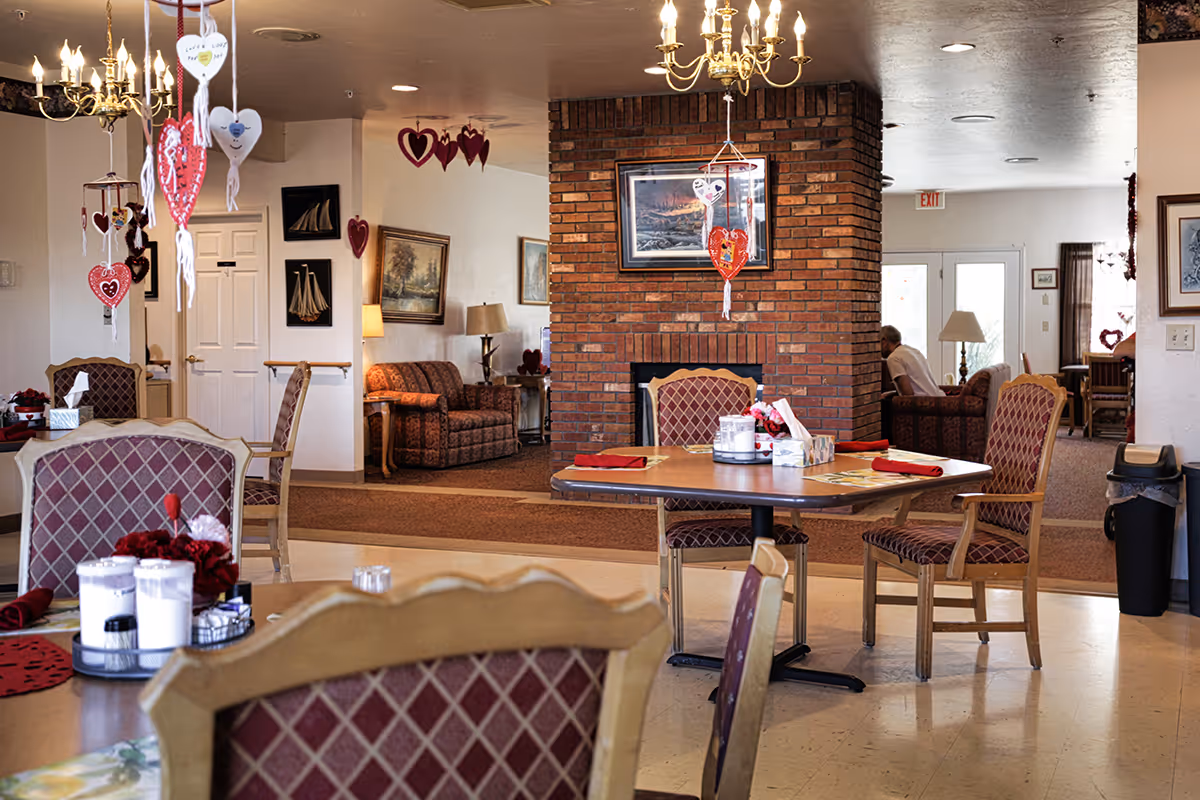 A cozy dining area in a senior living facility decorated with hanging heart ornaments. The room features tables with red napkins and floral centerpieces, upholstered chairs with a diamond pattern, and a brick fireplace with a framed picture above it. In the background, there are couches, lamps, and wall art, creating a warm and inviting atmosphere.